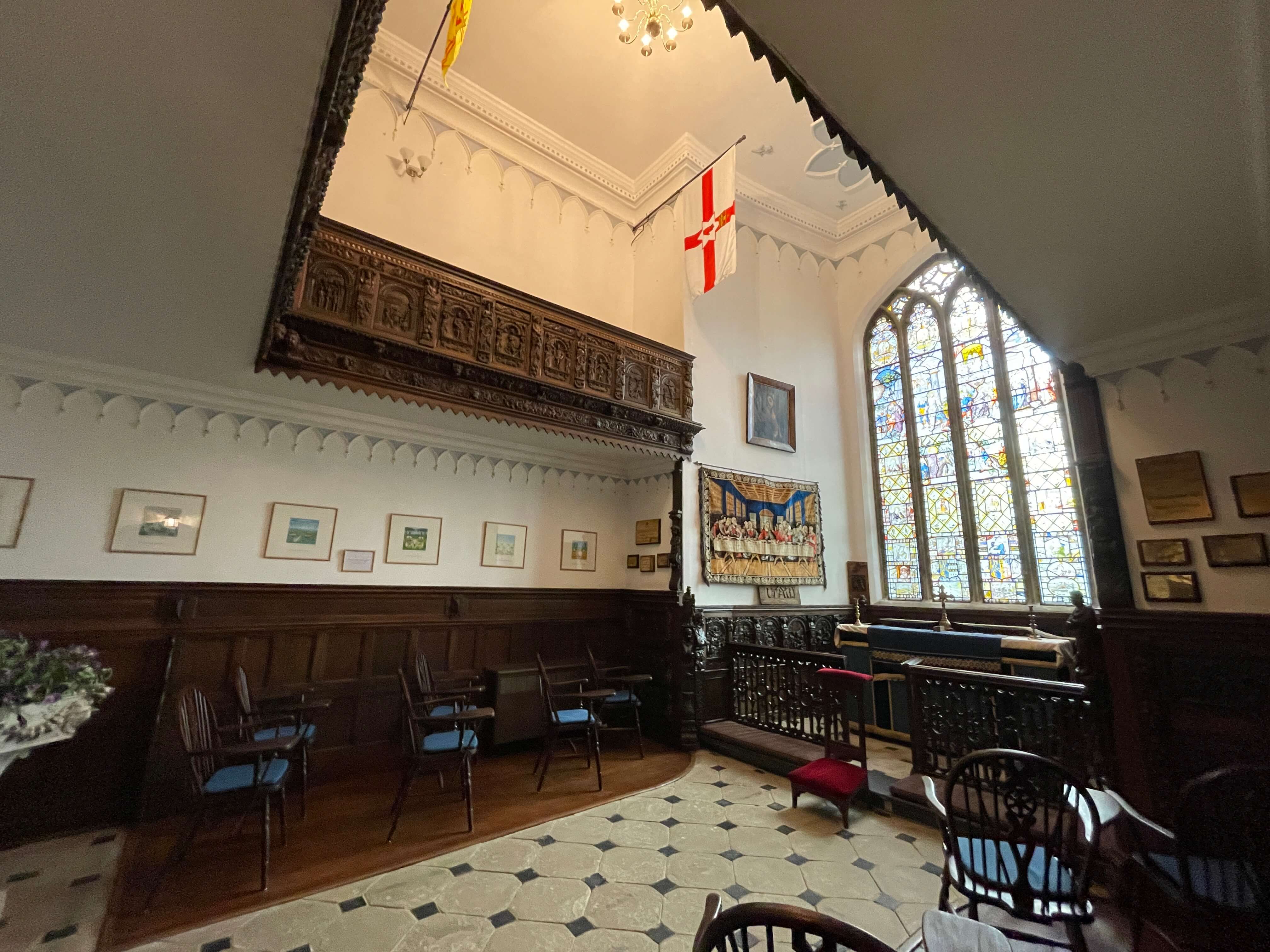 Historic chapel interior with carved wood paneling, stained glass window, St. George's flag, tapestry, and framed artwork on white walls.