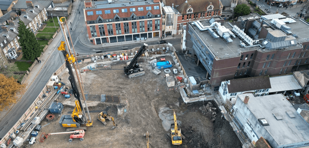 Aerial view of urban construction site with cranes and excavators surrounded by brick buildings and residential streets.