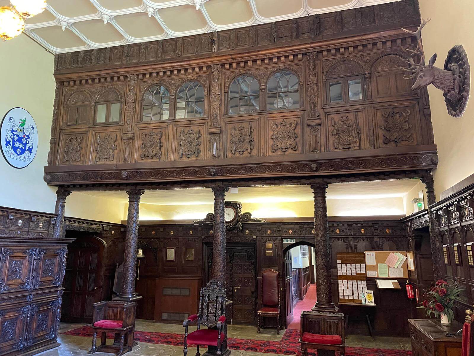 Grand historic hall with ornate dark wood carved paneling, a minstrels' gallery, stag head mount, coat of arms, and red upholstered chairs.