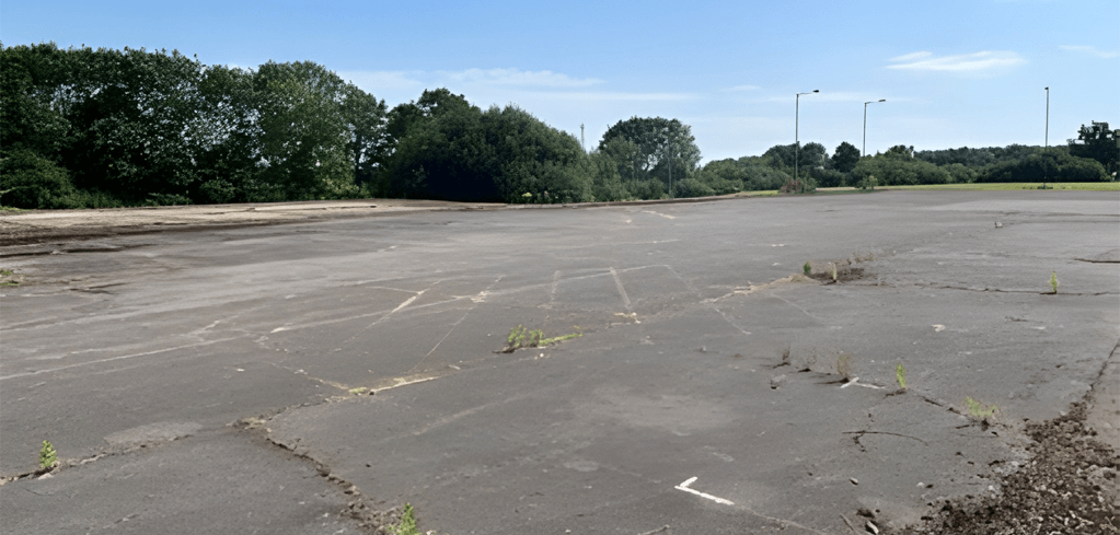 Empty cracked parking lot surrounded by green trees under blue sky, with weeds growing through pavement cracks.