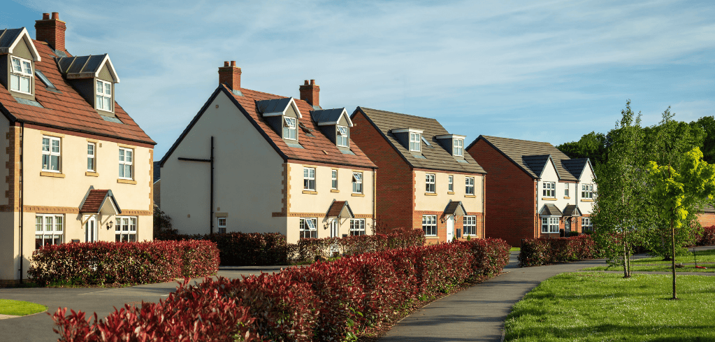 Row of modern suburban houses with red tile roofs, bordered by red hedges and green lawns under a blue sky.