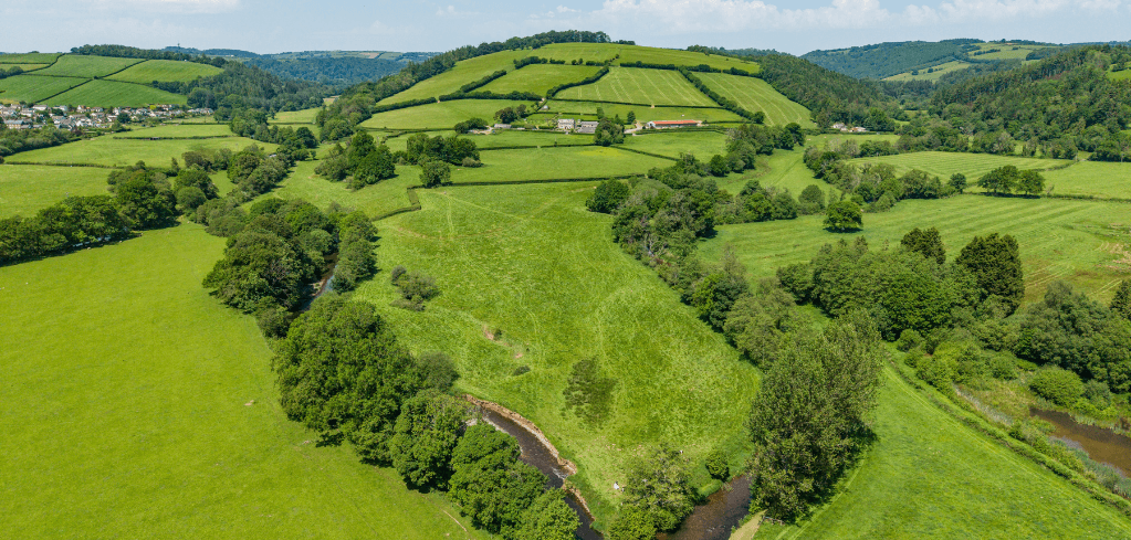 Aerial view of lush green rolling countryside with a winding river, farmland, trees, and a small village under a blue sky.