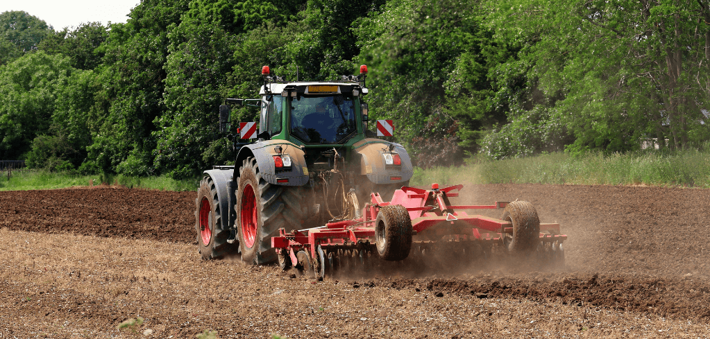 Green tractor pulling a red tilling attachment across a brown field, with lush trees in the background.