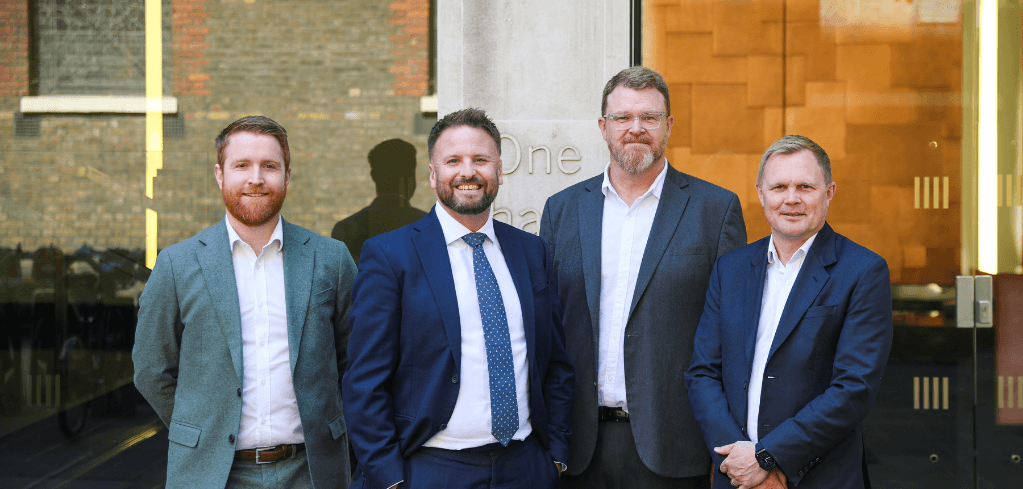 Four men in business attire smile while posing together outside a modern building with a brick facade.