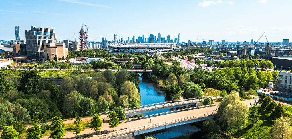 Aerial view of London's Olympic Park with lush greenery, a river, bridge, stadium, ArcelorMittal Orbit, and city skyline on a sunny day.