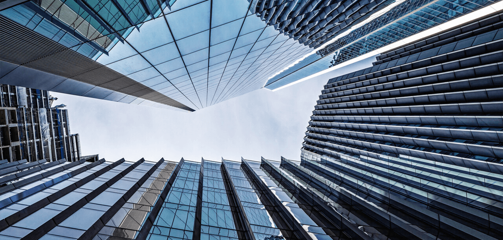 Looking up at modern glass skyscrapers from street level, with sleek blue facades converging toward a bright sky.