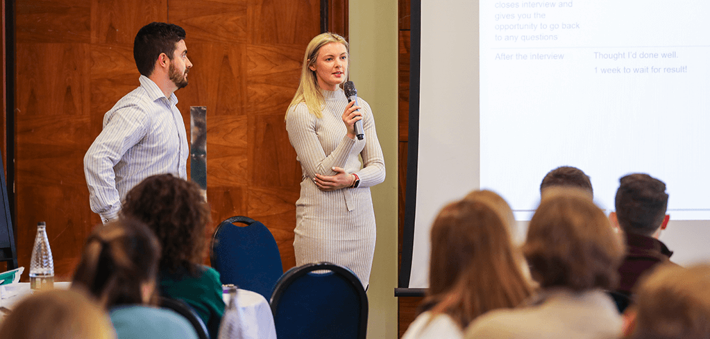 Two speakers presenting at a business conference, woman in cream dress holding microphone addressing seated audience.