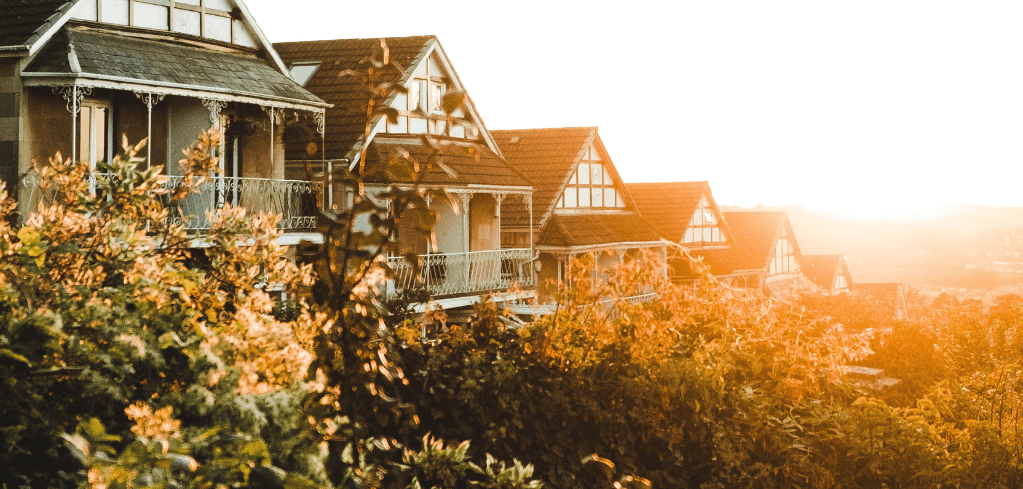 Row of Tudor-style houses with balconies bathed in golden sunset light, surrounded by autumn foliage on a hillside.
