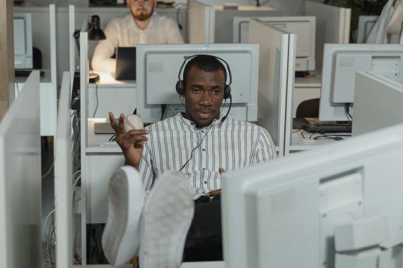 homem sentado num escritório apertado com os pés em cima da mesa a olhar para o monitor de um computador e com um headset e uma bola de tenis na mão. 