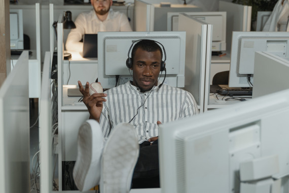 homem sentado num escritório apertado com os pés em cima da mesa a olhar para o monitor de um computador e com um headset e uma bola de tenis na mão.