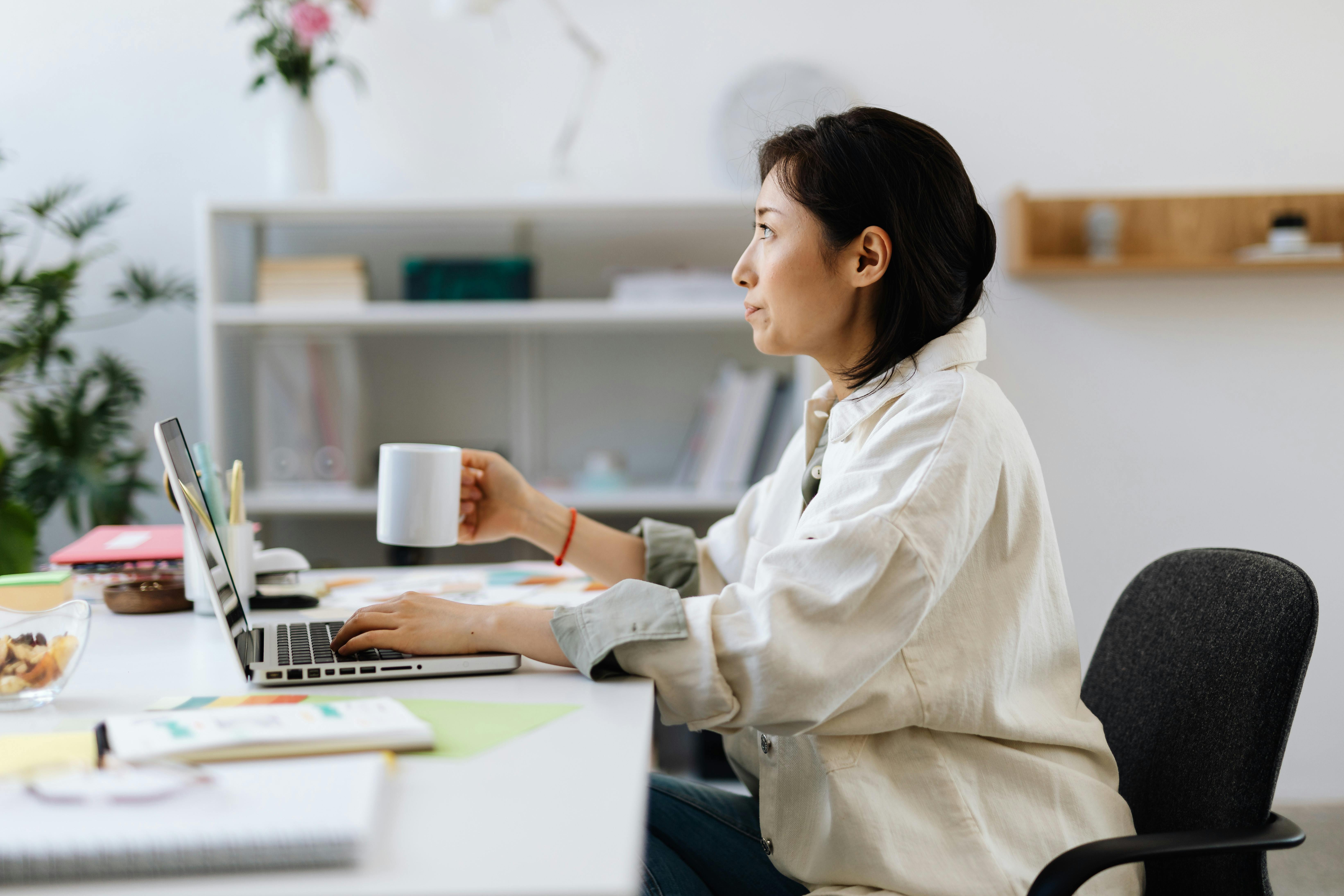Mulher sentada num escritório moderno, a trabalhar no portátil enquanto segura uma caneca, com um ambiente organizado e plantas decorativas ao fundo.