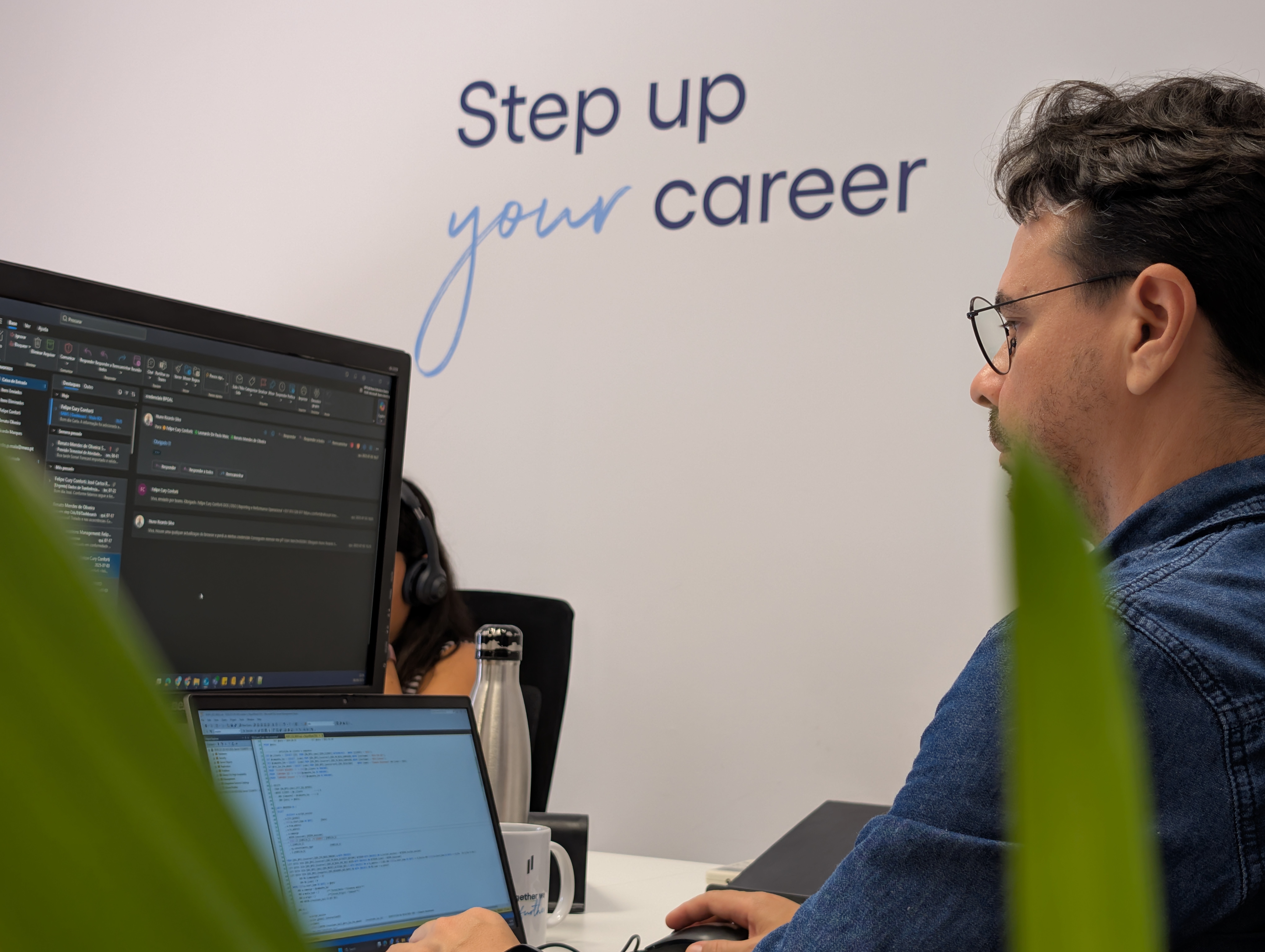 Colaboradores a trabalhar lado a lado em ambiente de escritório, com monitores e portáteis, sob uma parede com a frase motivacional ‘Step up your career
