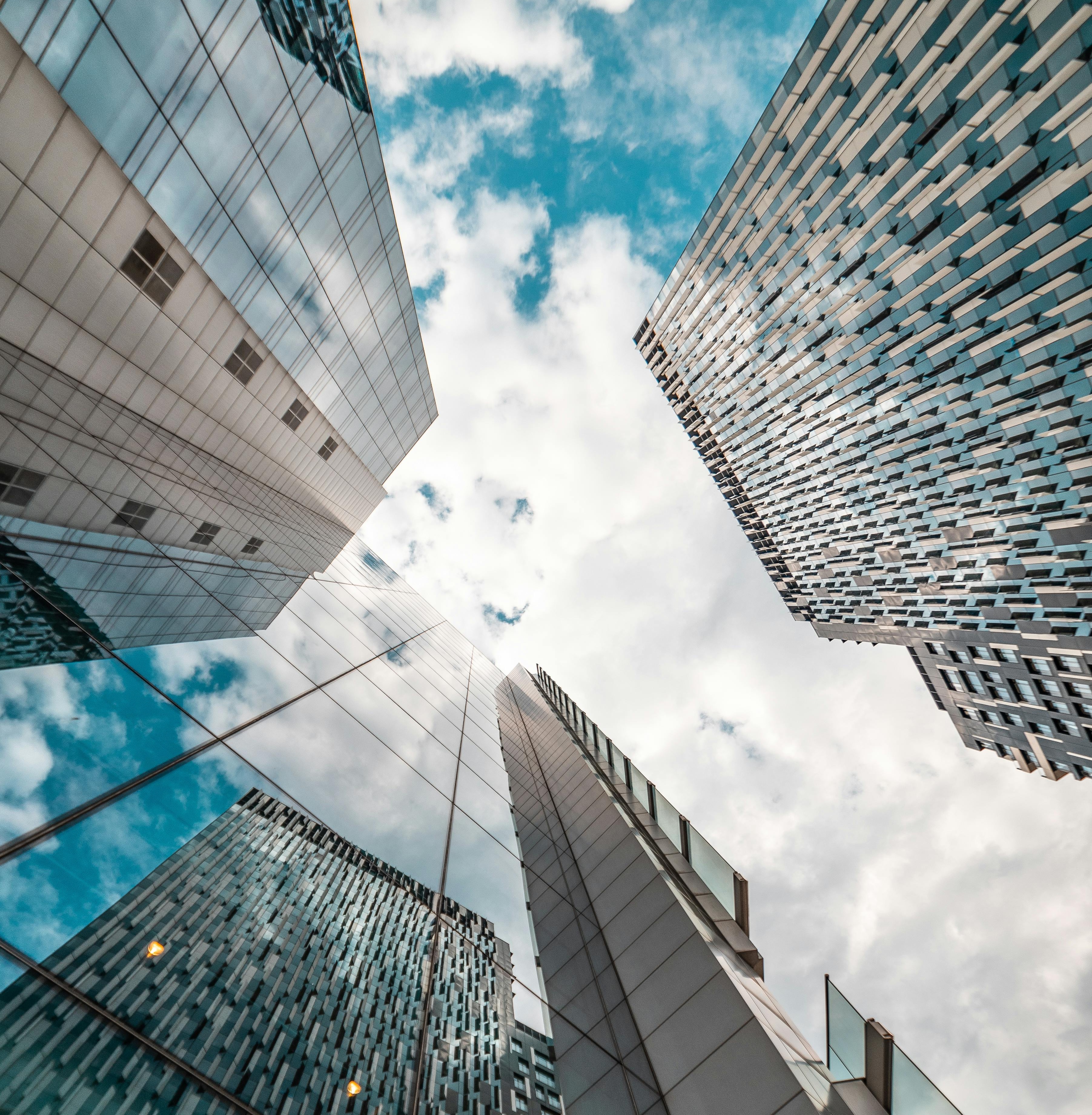 Perspetiva de baixo para cima de vários edifícios modernos envidraçados, refletindo o céu azul com nuvens, num ambiente urbano contemporâneo