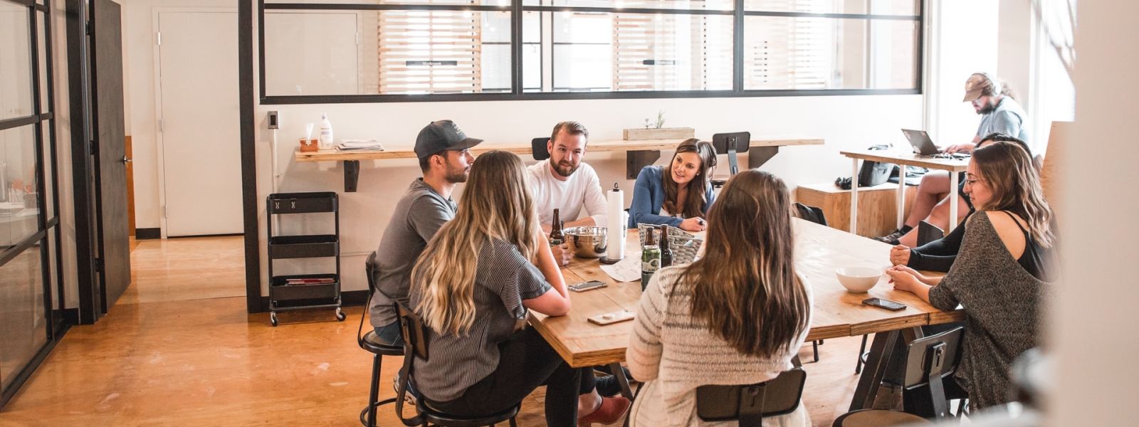 A group of 7 people sitting around an office table in discussion. There is someone in the background working with headphones on.