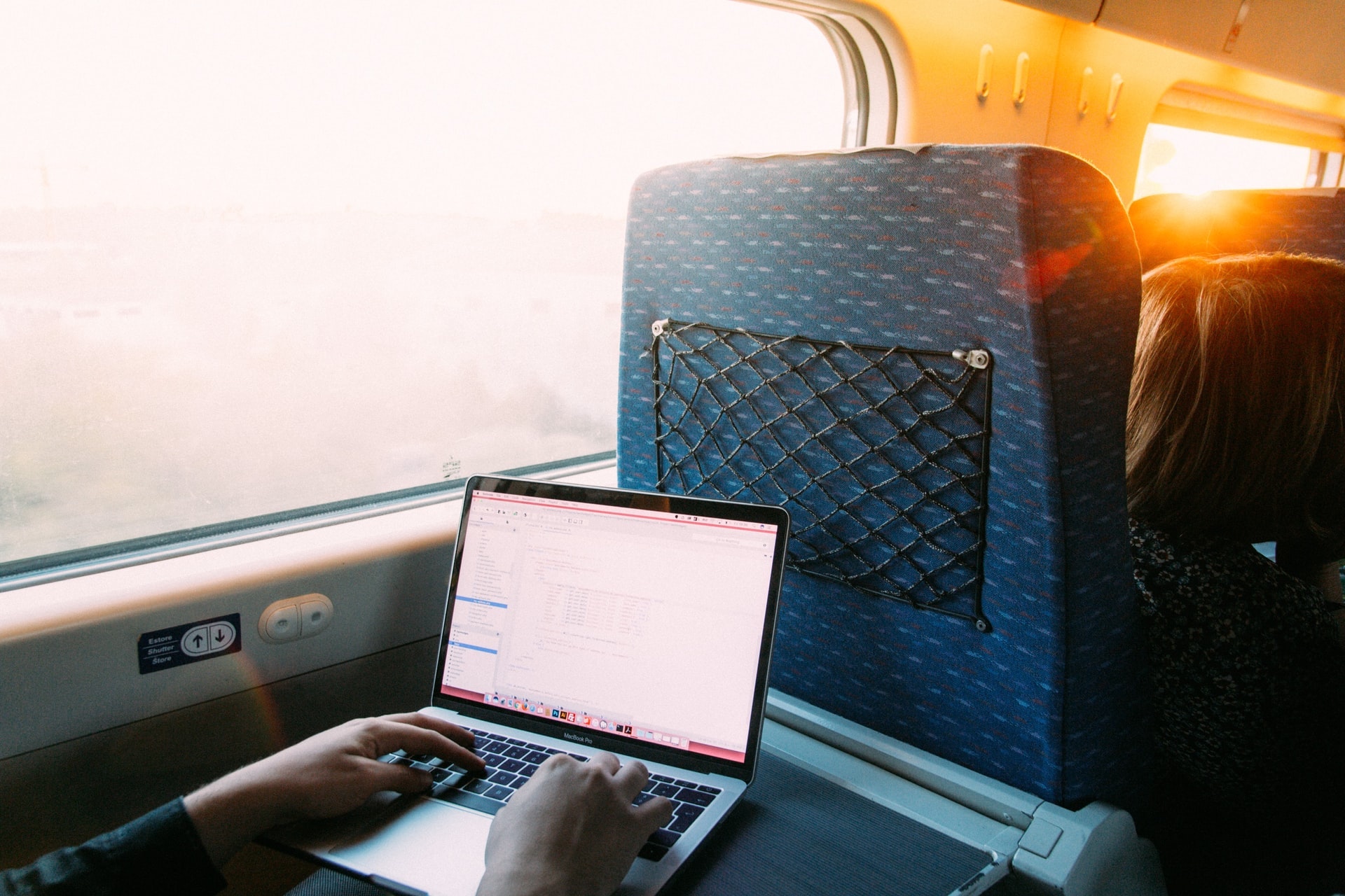 A laptop sitting on a tray table of a train, the sun is shining through the window and someone is typing on the keyboard.