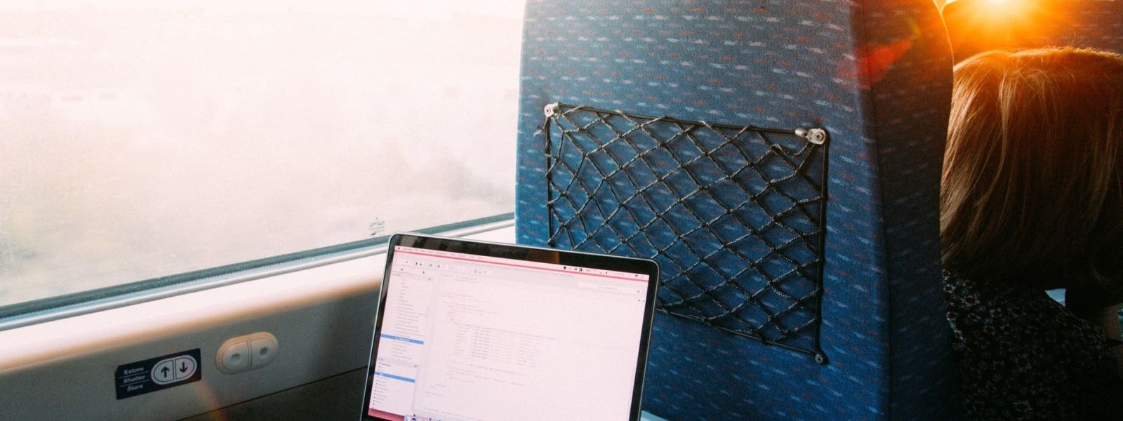A laptop sitting on a tray table of a train, the sun is shining through the window and someone is typing on the keyboard.