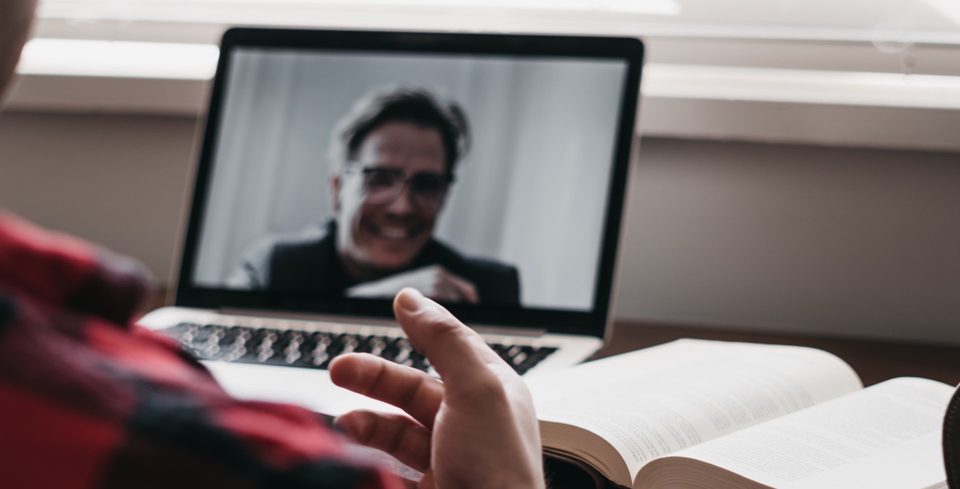 Someone is sitting at a desk with a laptop and a book open next to it. We can only see the person's shoulder and hand. On the laptop screen is an online call with someone with short hair and glasses.