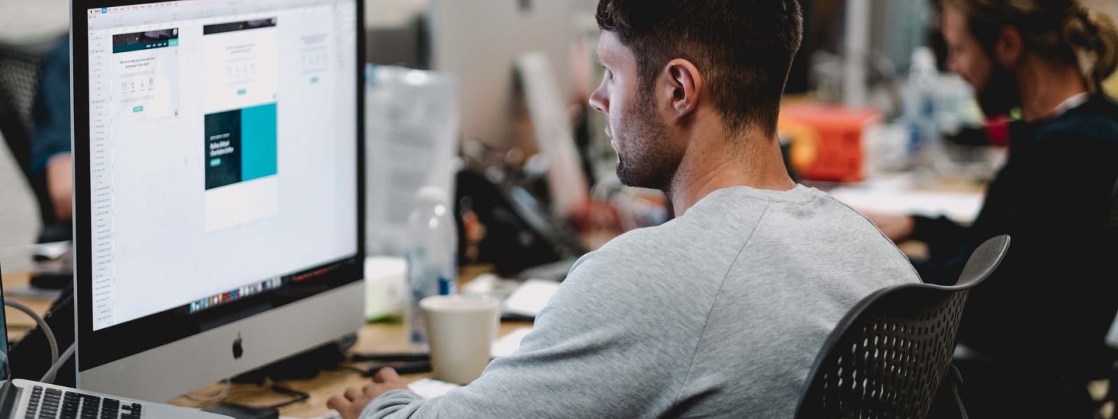 An image of a person working at a desktop Mac. There are others working in the background doing the same.