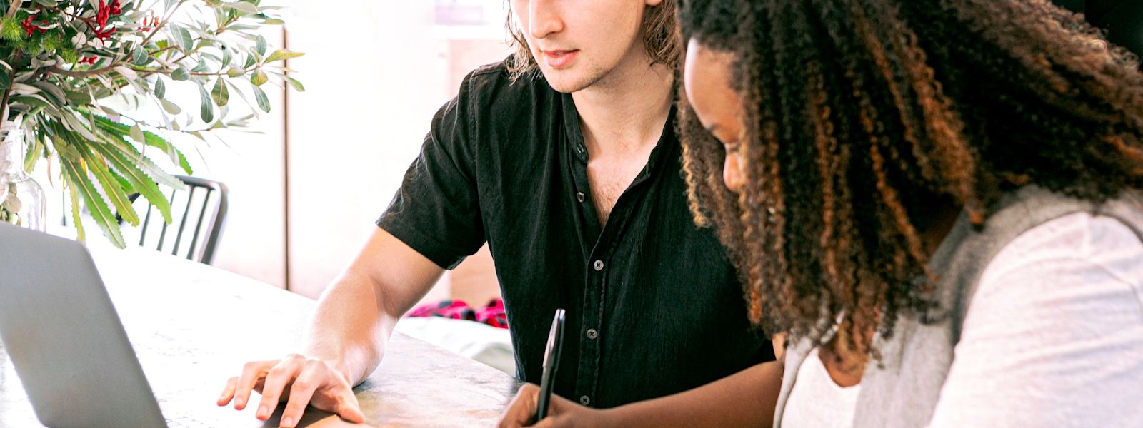 2 people sitting next to each other at a desk. The person on the left is looking at a laptop, while the person on the right is taking notes in a notebook.