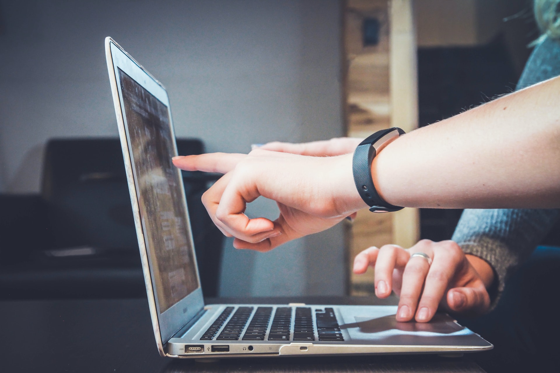 The side view of an Apple laptop with two people's hands pointing at the screen and using the trackpad.