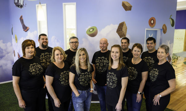 A group of 11 people wearing the same black tshirt, all standing in the Stem Rural office, in front of a wall with pictures of kiwifruit, hay bales and work boots.