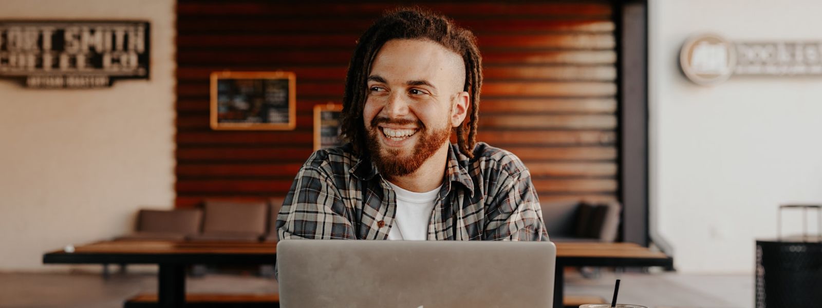 A person is sitting at a table with their laptop in front of them. They're looking to their right and smiling. There's an ice coffee next to them, and they have orange long hair and an orange trimmed beard.