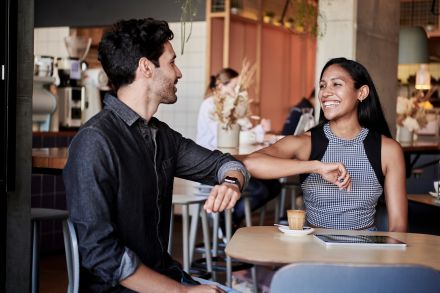 Two people at a cafe, bumping elbows and smiling.