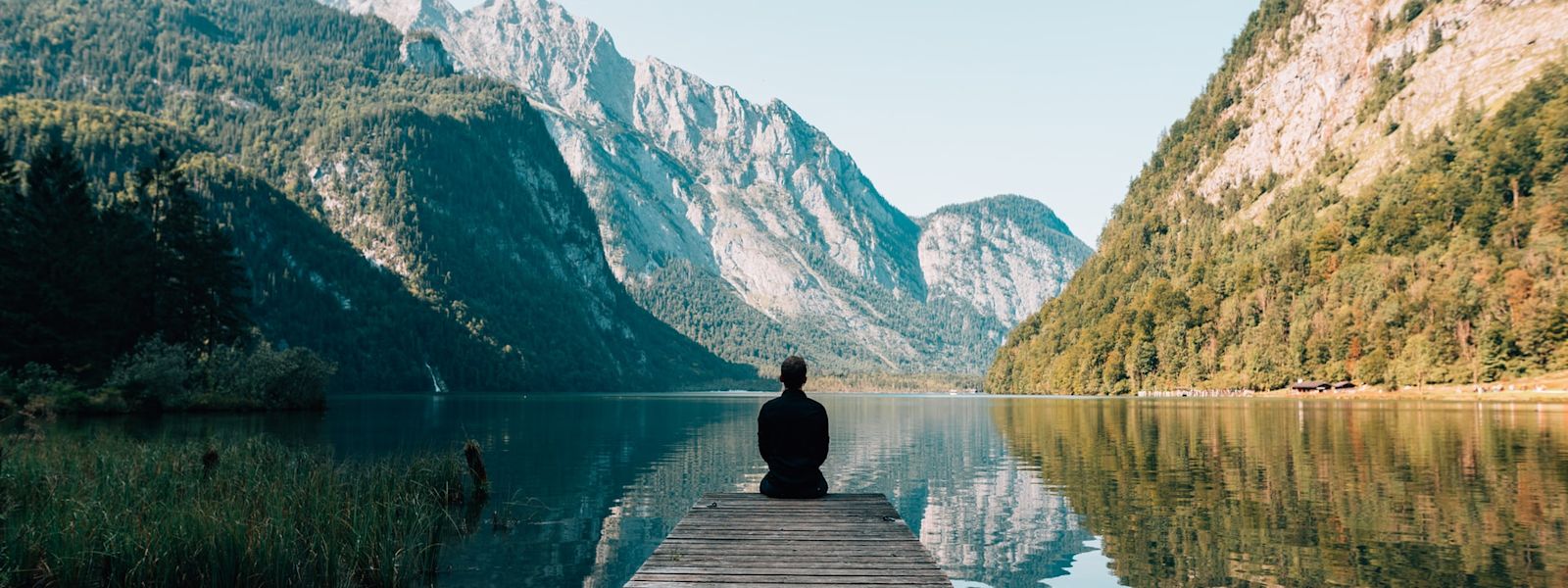 A person sitting at the end of a jetty, looking out onto a lake with towering mountains on both sides. In the distance, there are snow-capped mountains, and the lake is reflecting the mountains and pale blue sky.