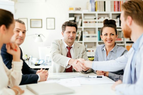 A group of professionals in a modern office setting, with two individuals shaking hands across a table, symbolizing partnership or merger discussions in the accounting industry.