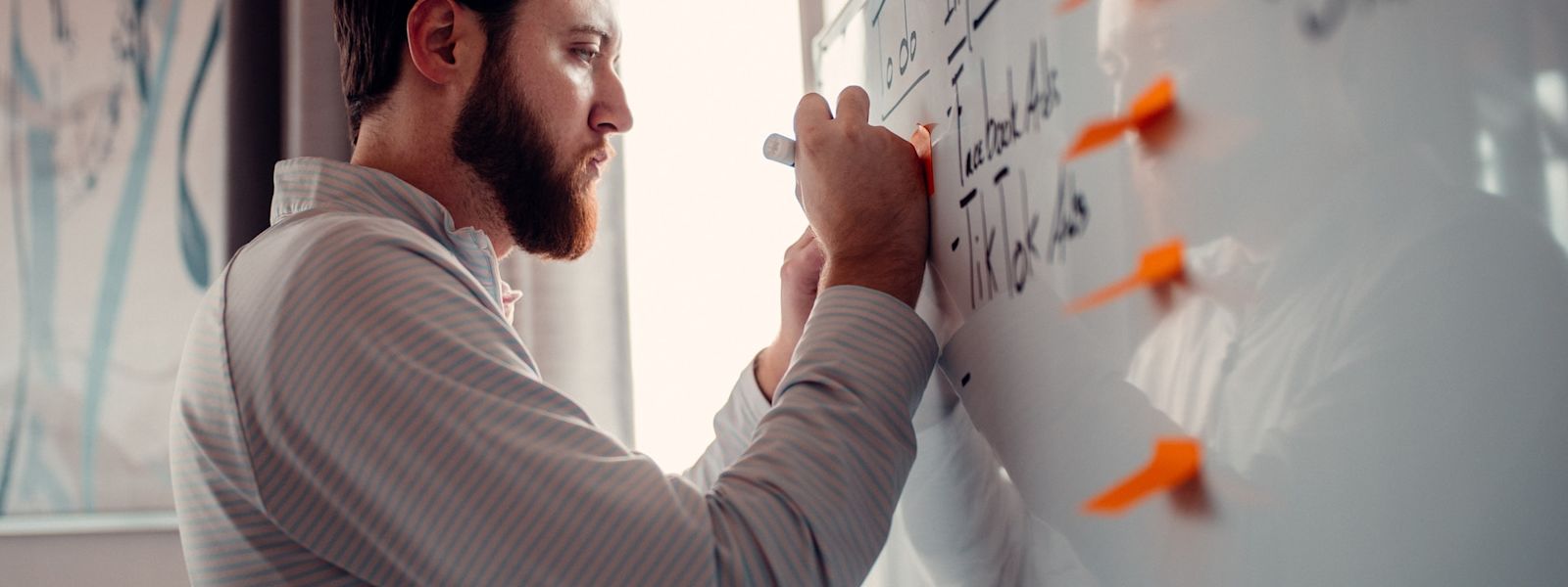 A person is writing on a whiteboard that is covered in writing and 4 orange sticky notes.