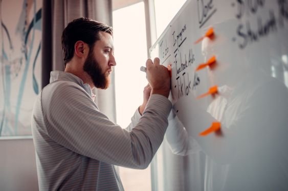 A person is writing on a whiteboard that is covered in writing and 4 orange sticky notes.