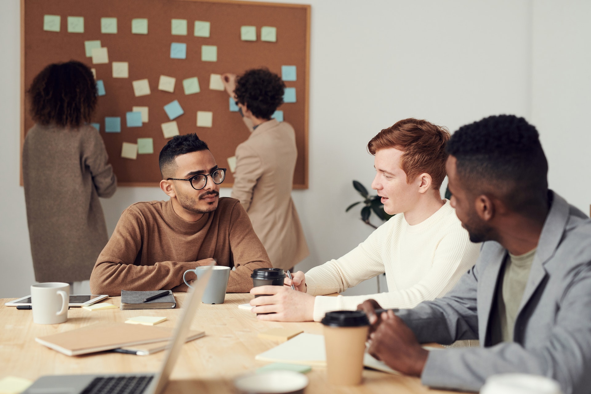 3 people sitting around a desk making business decisions, with 2 others standing behind them in front of a pinboard covered in sticky notes.
