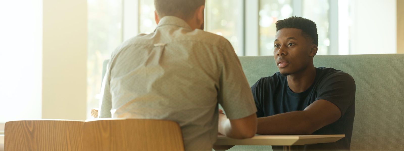 Two people sitting opposite each other at a table. One person has their back to the camera and is wearing a light shirt, the other person is facing the camera and is wearing a dark tshirt.