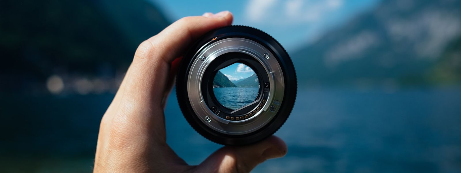 Someone is holding a camera lens up to two mountains meeting at a valley. The rest of the image is blurry, apart from the person's hand, the lens, and the image within the lens, which are the mountains meeting.