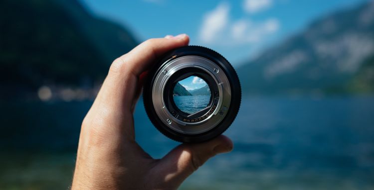 Someone is holding a camera lens up to two mountains meeting at a valley. The rest of the image is blurry, apart from the person's hand, the lens, and the image within the lens, which are the mountains meeting.