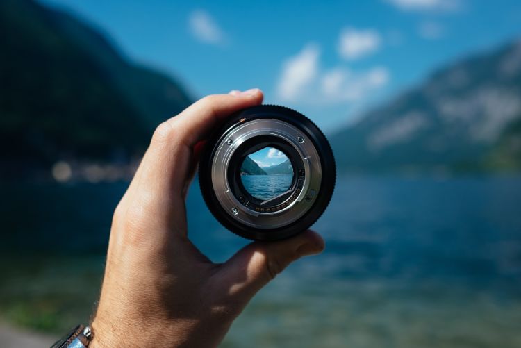 Someone is holding a camera lens up to two mountains meeting at a valley. The rest of the image is blurry, apart from the person's hand, the lens, and the image within the lens, which are the mountains meeting.