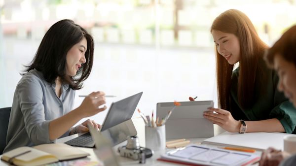 Two accounting professionals collaborating at a desk, one working on a laptop and the other on a tablet, surrounded by office supplies. Their interaction reflects effective capacity planning and teamwork.