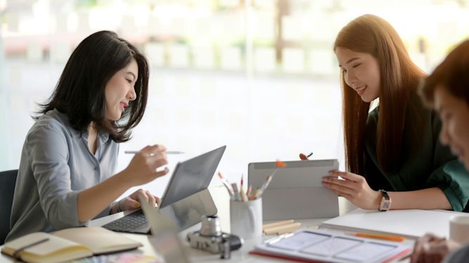 Two accounting professionals collaborating at a desk, one working on a laptop and the other on a tablet, surrounded by office supplies. Their interaction reflects effective capacity planning and teamwork.