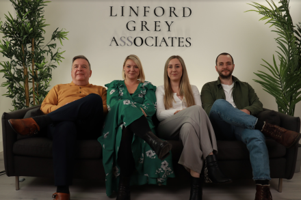 The Linford Grey team sitting next to each other on a sofa, legs crossed, slightly smiling.