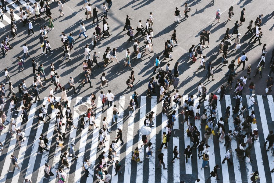 An aerial view of a busy crosswalk. People are walking in every direction, spilling out over the crossing road markings.
