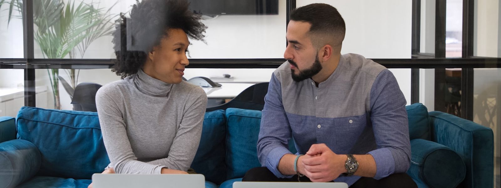 Two people sitting next to each other on a sofa in discussion, with their laptops on the table in front of them.