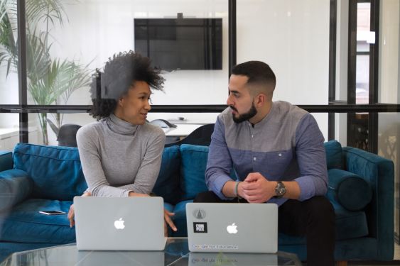 Two people sitting next to each other on a sofa in discussion, with their laptops on the table in front of them.