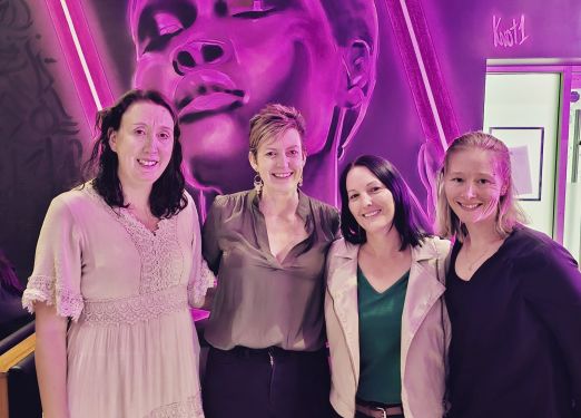 Sarah Stein, owner of Miss Efficiency, and some members of her team. They're at a welcome dinner for new team member, Kelly, and are standing in front of a purple and grey mural of a person.