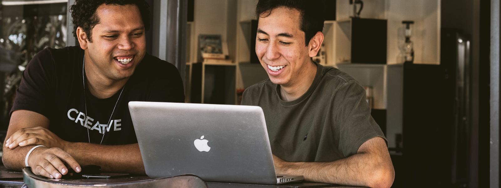 Two people sitting at a table and smiling at a laptop screen in front of them.