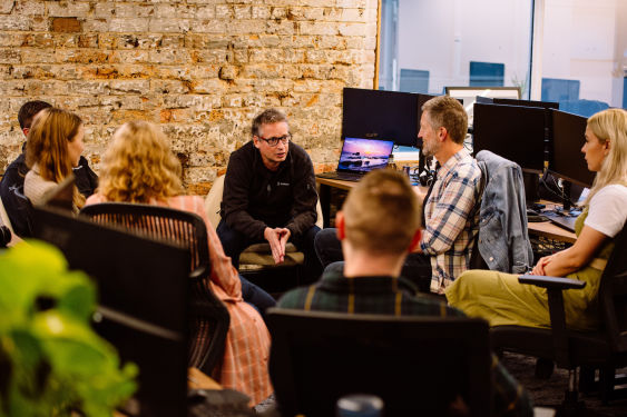 Stuart McLeod, Karbon CEO and Co-Founder, sitting with 6 other Karbon staff members. They're in deep discussion in their office. There are monitors, they're sitting in office chairs, and there's a brown exposed brick feature wall that makes up most of the background.