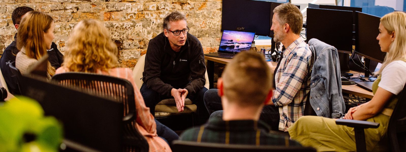 Stuart McLeod, Karbon CEO and Co-Founder, sitting with 6 other Karbon staff members. They're in deep discussion in their office. There are monitors, they're sitting in office chairs, and there's a brown exposed brick feature wall that makes up most of the background.