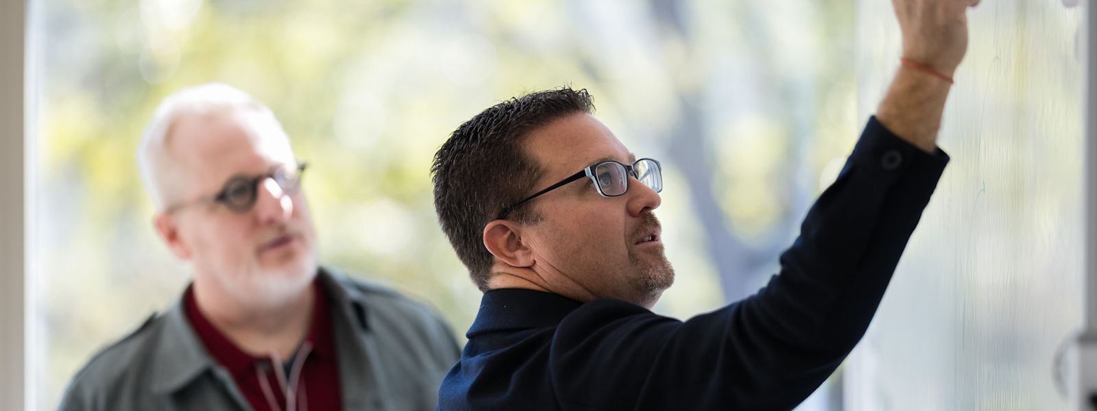 Ian Vacin and Jason Blumer stand at a whiteboard with Ian writing while Jason watches attentively during a planning discussion in a bright office.