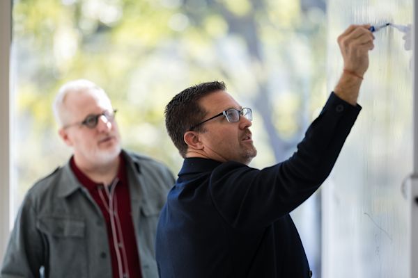 Ian Vacin and Jason Blumer stand at a whiteboard with Ian writing while Jason watches attentively during a planning discussion in a bright office.