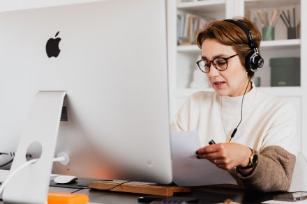 A person is working behind a desktop Mac. They're wearing a phone headset and they're holding a sheet of paper. It looks like they're on a call with a client.