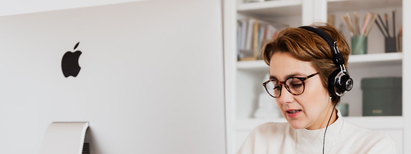 A person is sitting at a desktop Mac computer. They're wearing glasses, and a mic/ headphone headset, and they're looking at some paperwork.